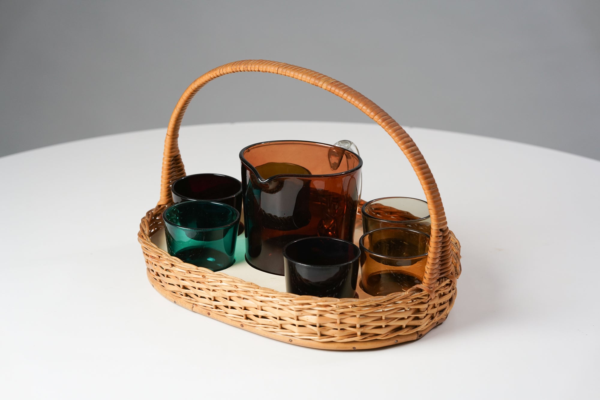 A set that includes six glass cups,a glass ewer and waved basket. The cups are in following colors: Turquoise, black, green, purplish red, yellow and brownish. The ewer is dark orange.