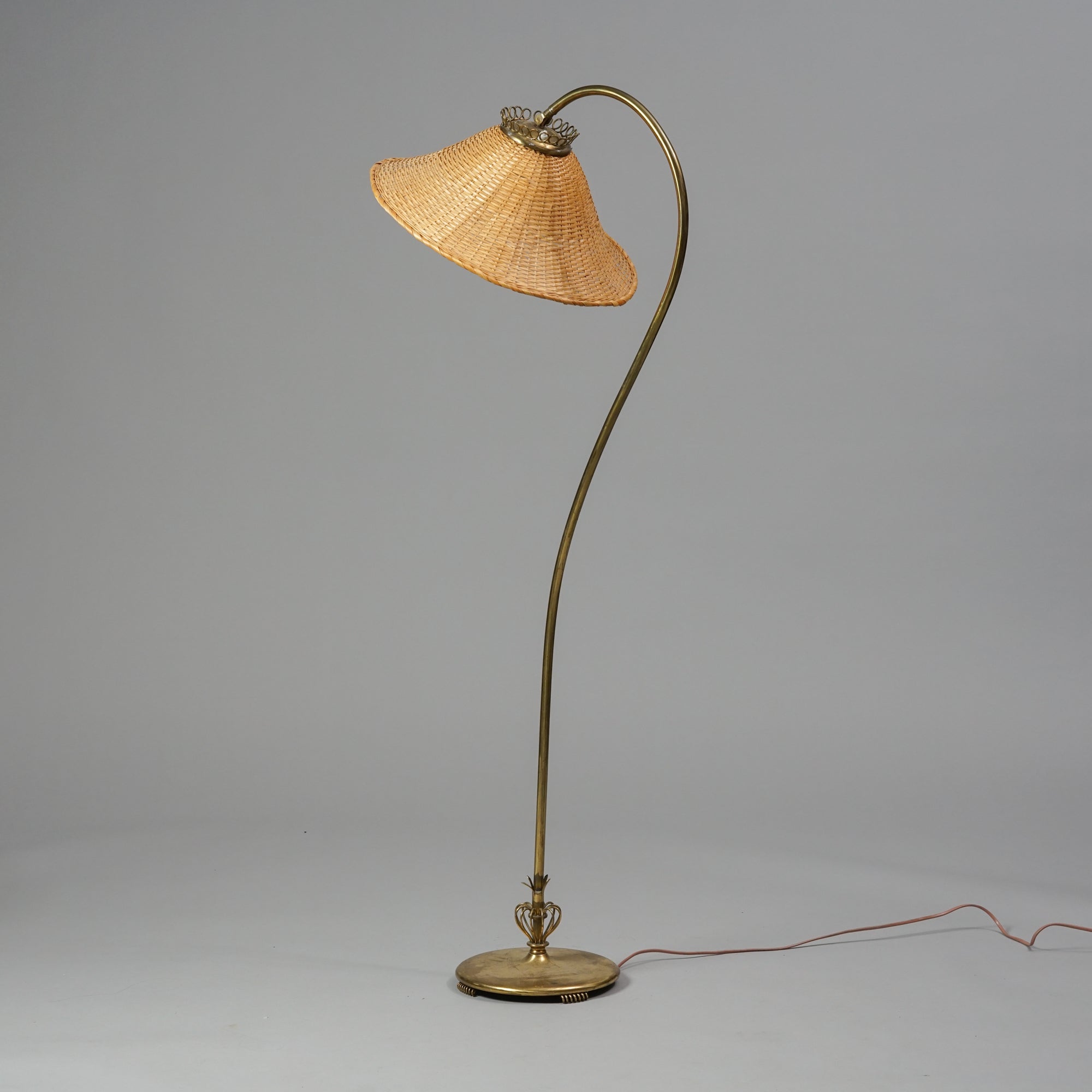 A brass lamp with decorations at the base. The leg is thin and curved, while the shade is made of basket.