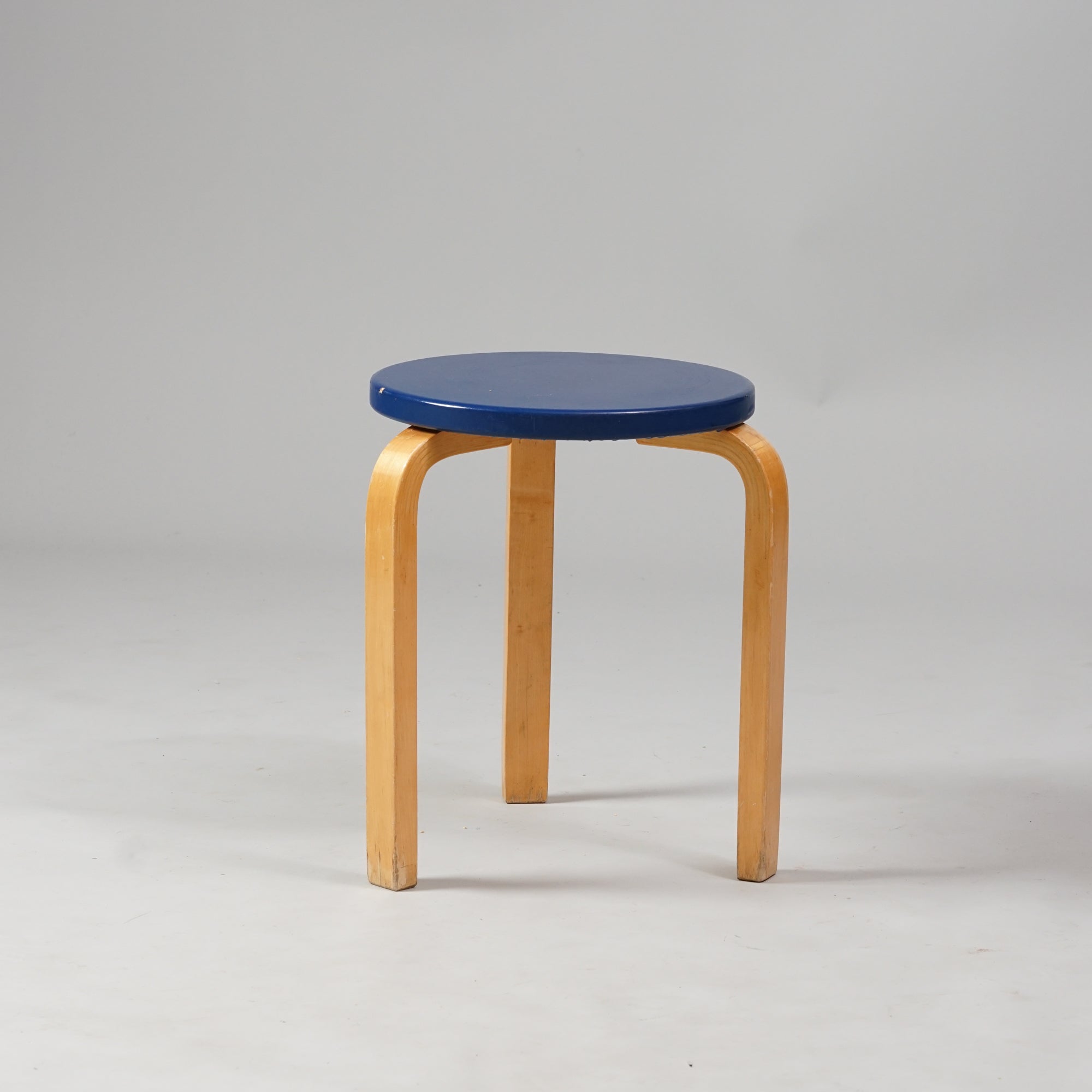 Three legged stool with a round blue seat. The stool is made of birch.